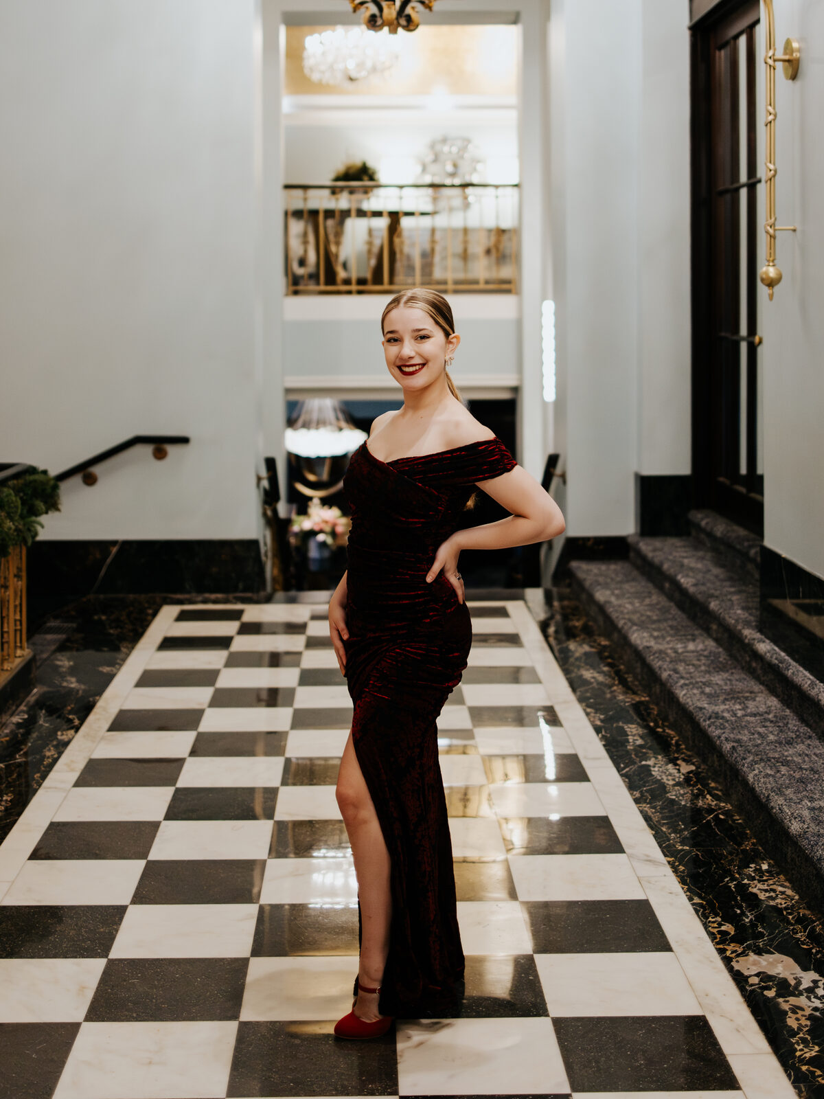 Ariella Sinclair in a burgundy velvet off-shoulder gown standing on a checkered marble floor beneath a grand chandelier