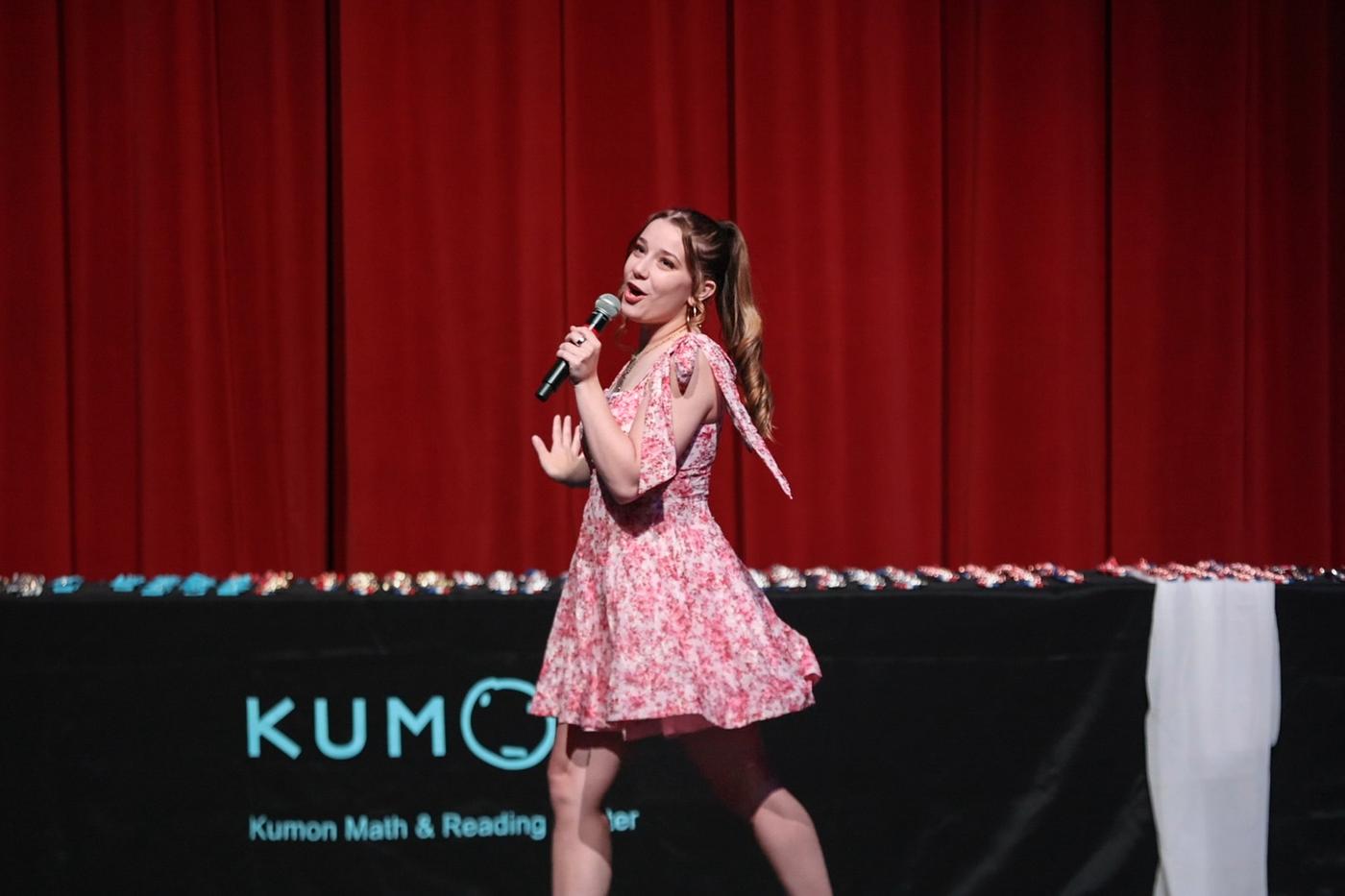 Ariella Sinclair singing on stage in a pink floral dress with dynamic microphone pose